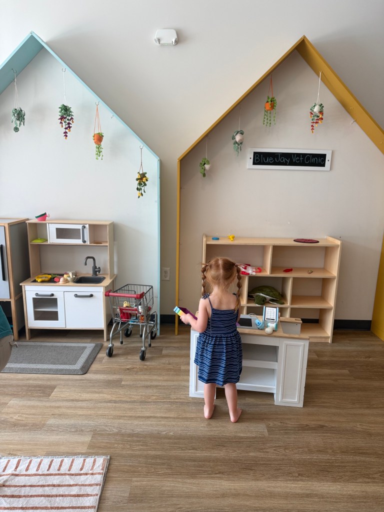 A young girl in a striped blue dress plays at a pretend Blue Jay Vet Clinic in a bright Montessori-style indoor play space with toy kitchens, shelves, and hanging plants.